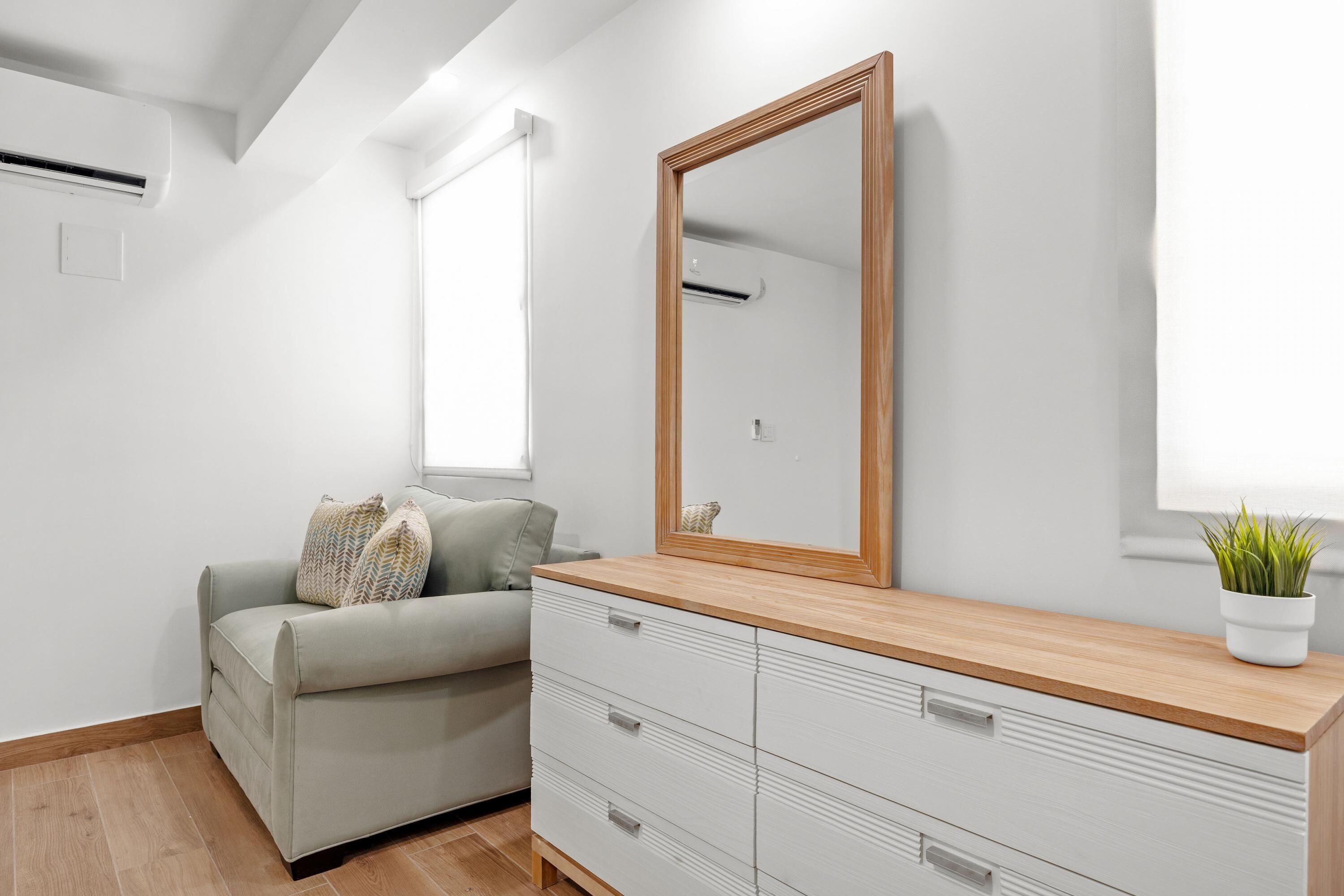 Una camera da letto moderna con un divano grigio, una cassettiera in legno, un grande specchio e una pianta in vaso.