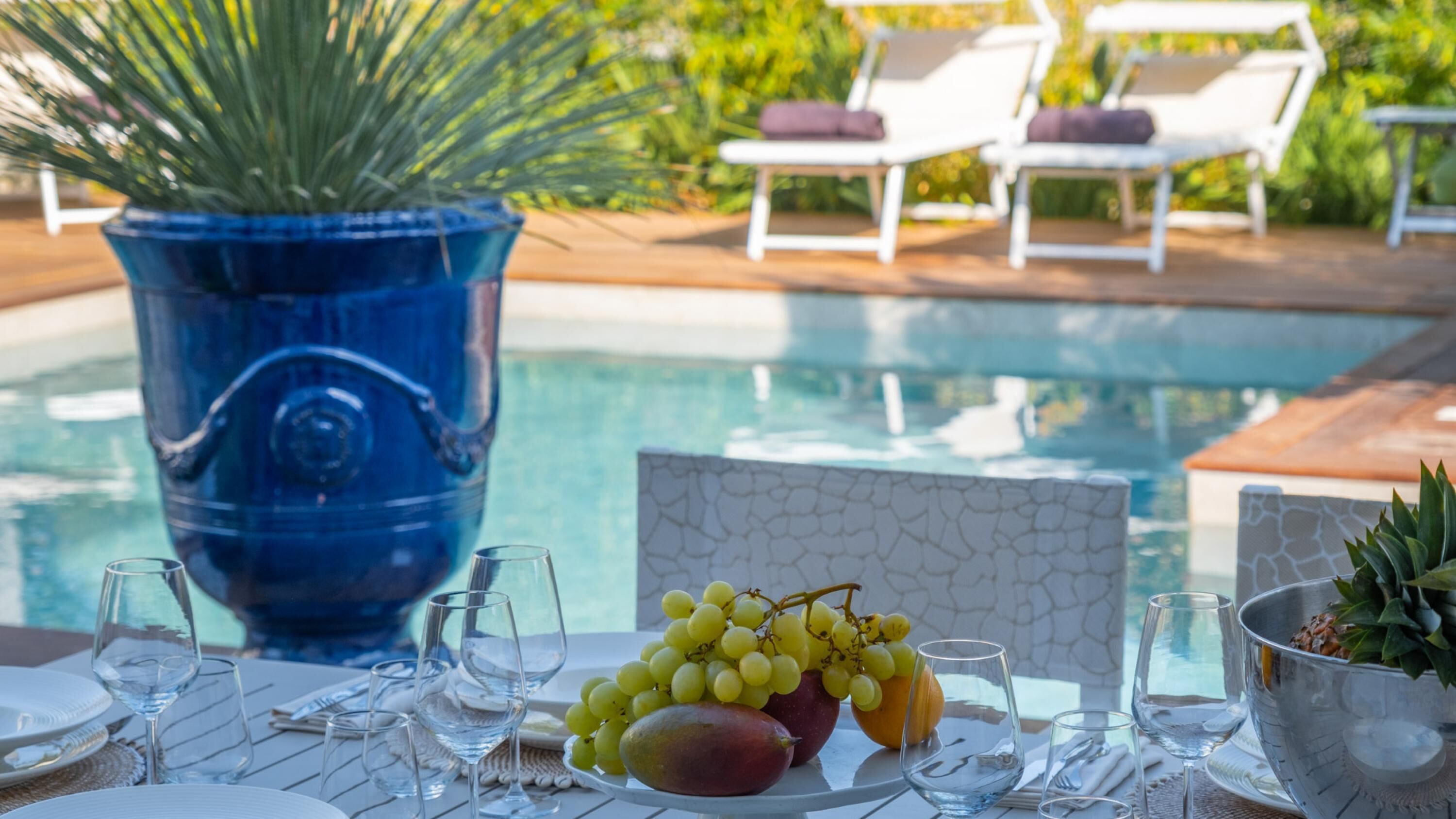 A poolside table with glasses, a fruit bowl, and a decorative vase.