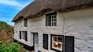 A traditional thatched-roof house with white walls and black shutters, featuring a small window with a visible interior light. - Lewcott (Sennen)