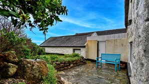 A stone-paved courtyard with a blue bench, a stone wall, and a building with a grey roof. - Lewcott (Sennen)