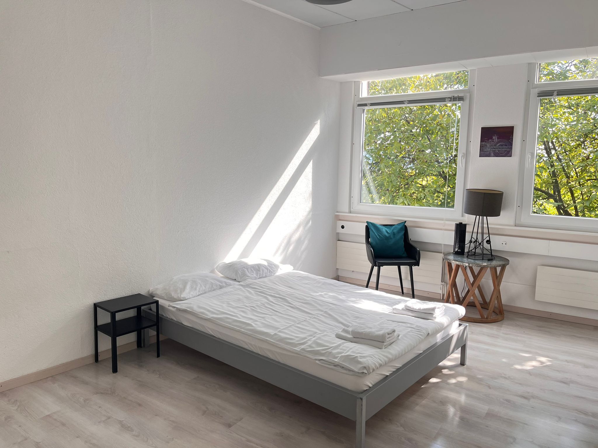 Ein modernes Schlafzimmer mit einem Bett, einem kleinen Tisch, einem Stuhl und einer Lampe. Große Fenster bieten Blick auf Bäume.