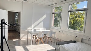 A modern, well-lit room with a dining table, chairs, a bed, and large windows offering a view of greenery. - Bright, comfy, industrial looking (Fällanden)