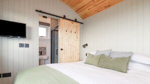 A bedroom with a bed, a television mounted on the wall, a wooden ceiling, and a sliding door leading to a bathroom. - Northcott Hut (Bude)
