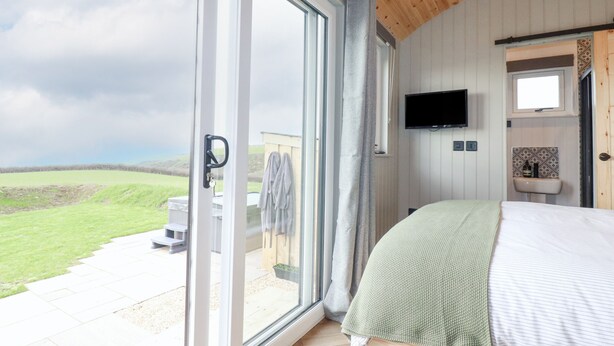 A bedroom with a large sliding glass door leading to a balcony, a television mounted on the wall, and a small sink. - Northcott Hut (Bude)
