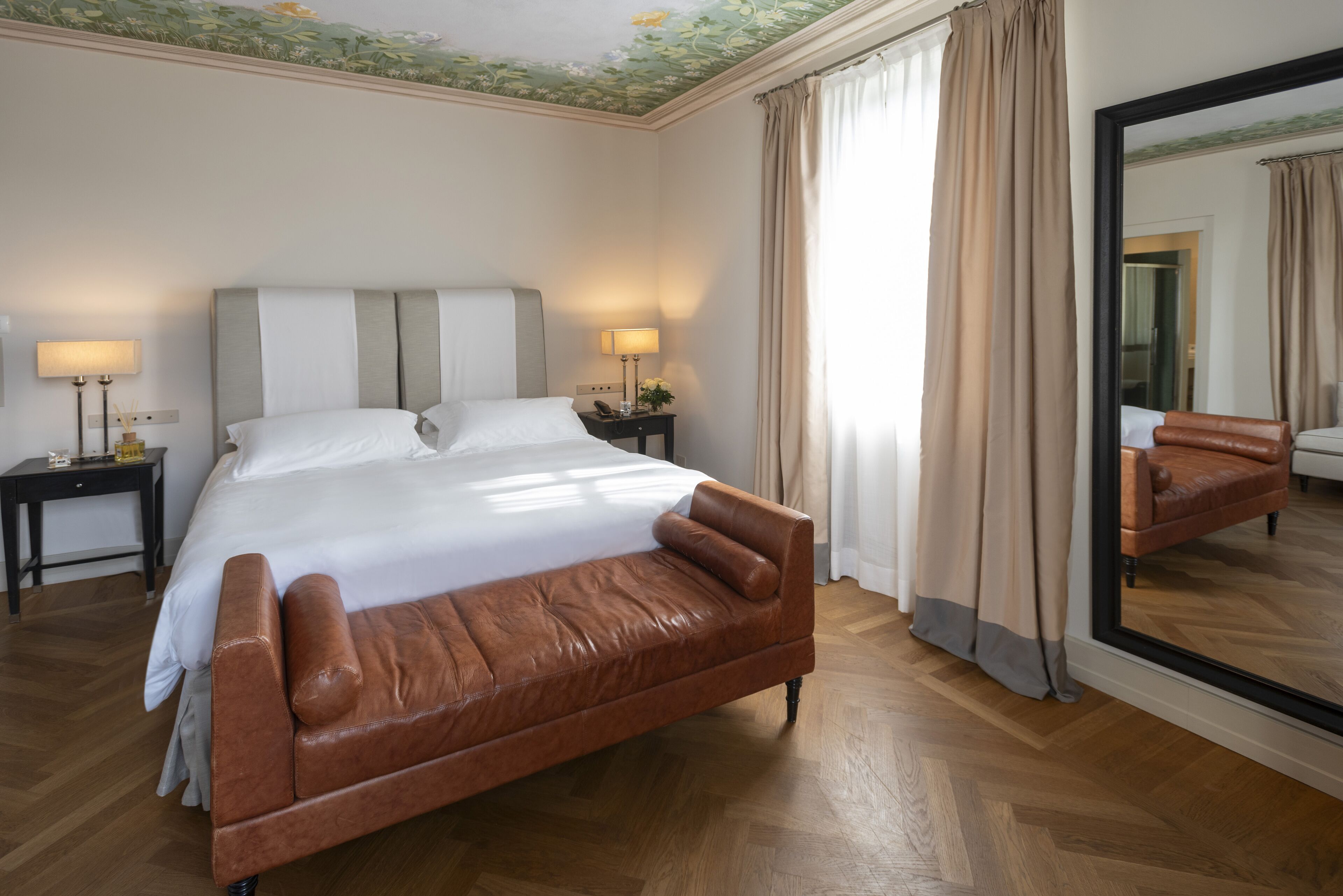 A hotel room with a large bed, two bedside tables with lamps, a large mirror, and a leather sofa.