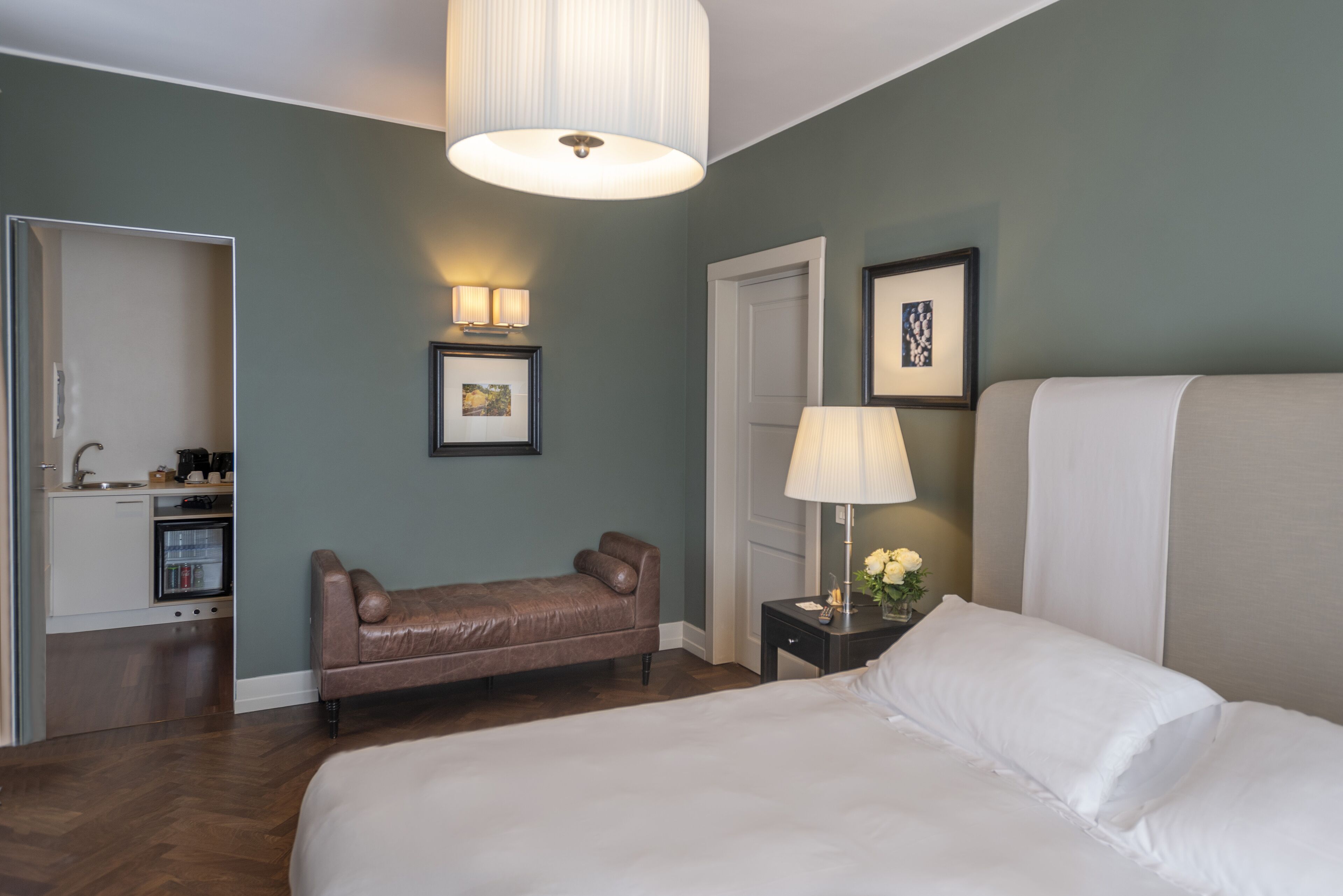 A hotel room with a large bed, a small sofa, a bedside table with a lamp, a picture frame, and a door leading to a kitchenette.