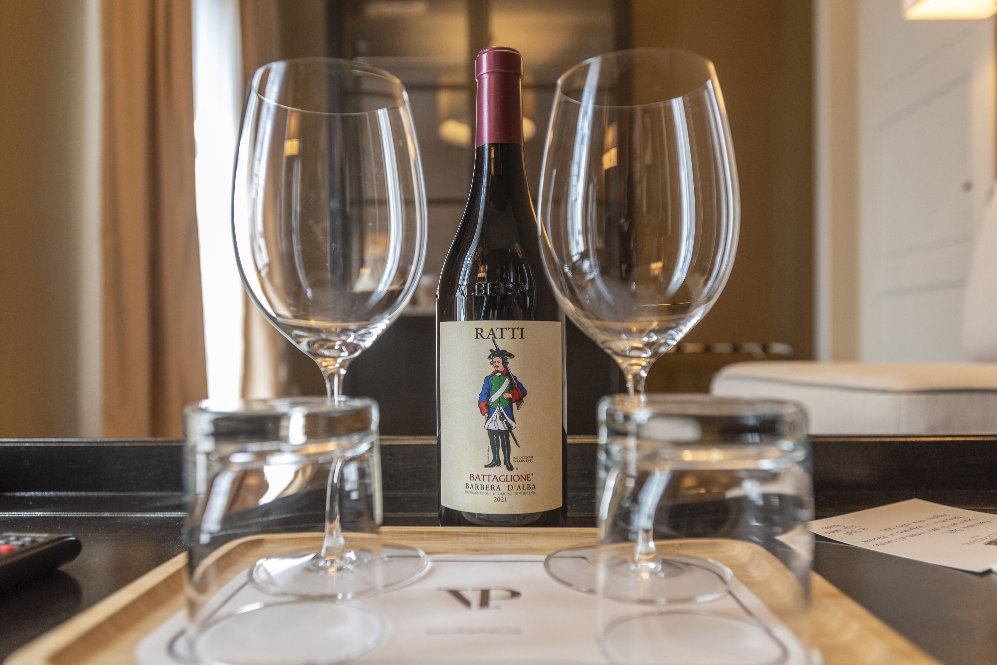 A bottle of Battagione Barbera d'Alba wine with two empty wine glasses on a tray.