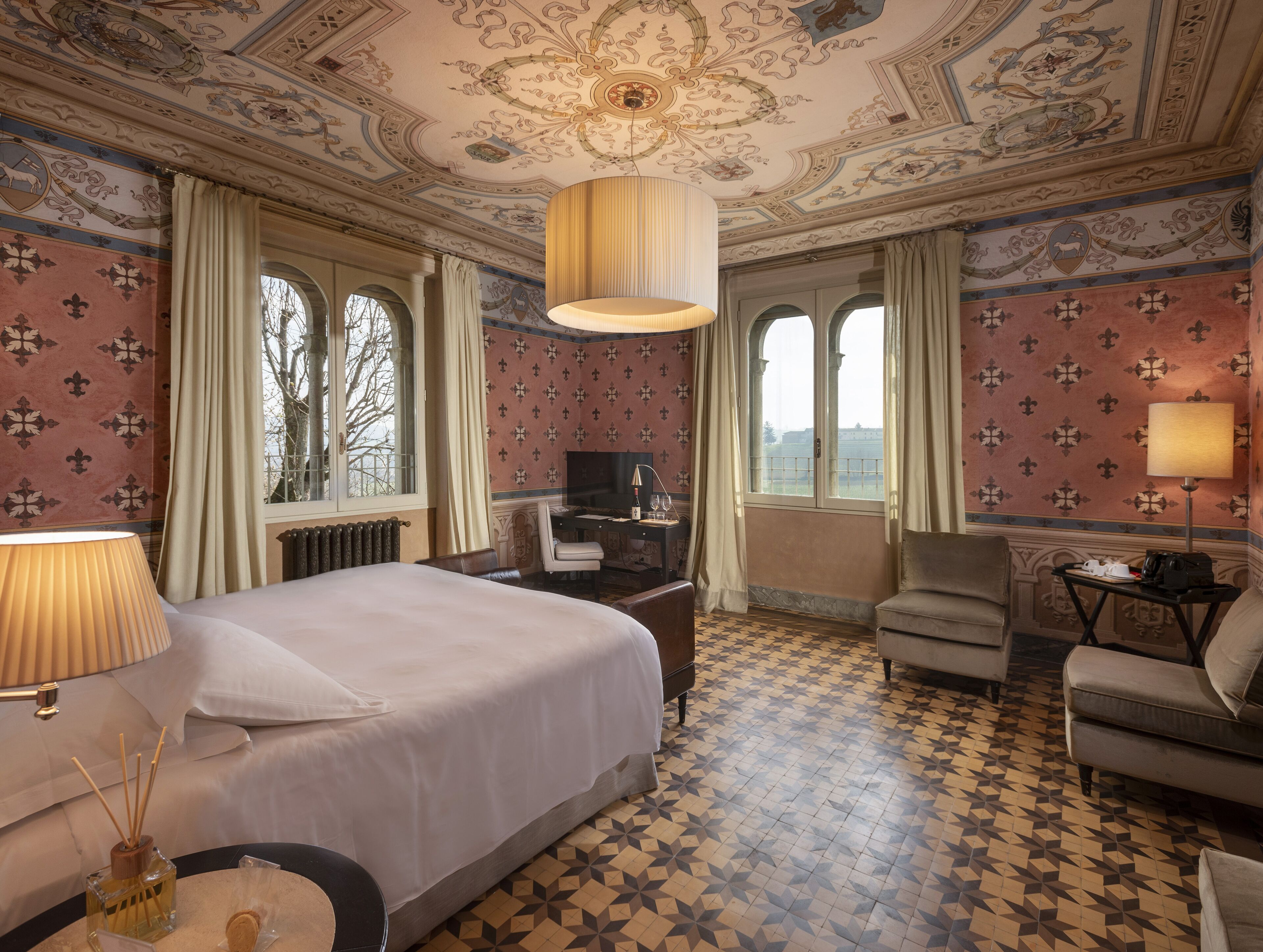 A hotel room with a large bed, a desk with a computer, a chair, and a sofa. There are two windows with curtains, and the walls have a patterned wallpaper.