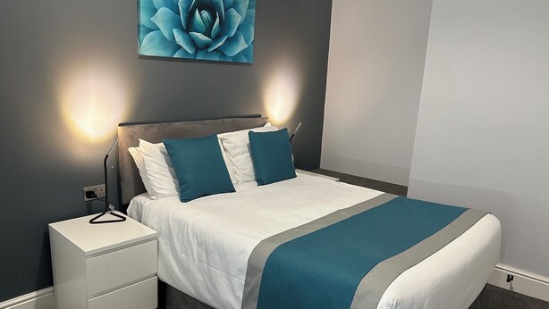 A neatly made bed with white and teal bedding, a grey headboard, and a bedside table with a lamp. - Arlan House Hotel (Basingstoke)