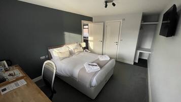A hotel room with a bed, bedside table, and a desk with a chair. There is a wardrobe and a television mounted on the wall.