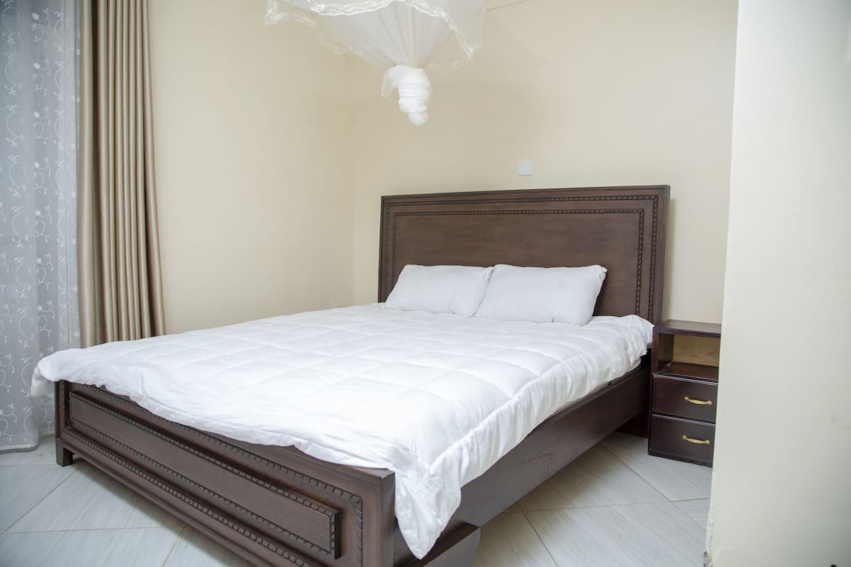 A bedroom with a wooden bed, white bedding, a nightstand with drawers, and a sheer curtain.