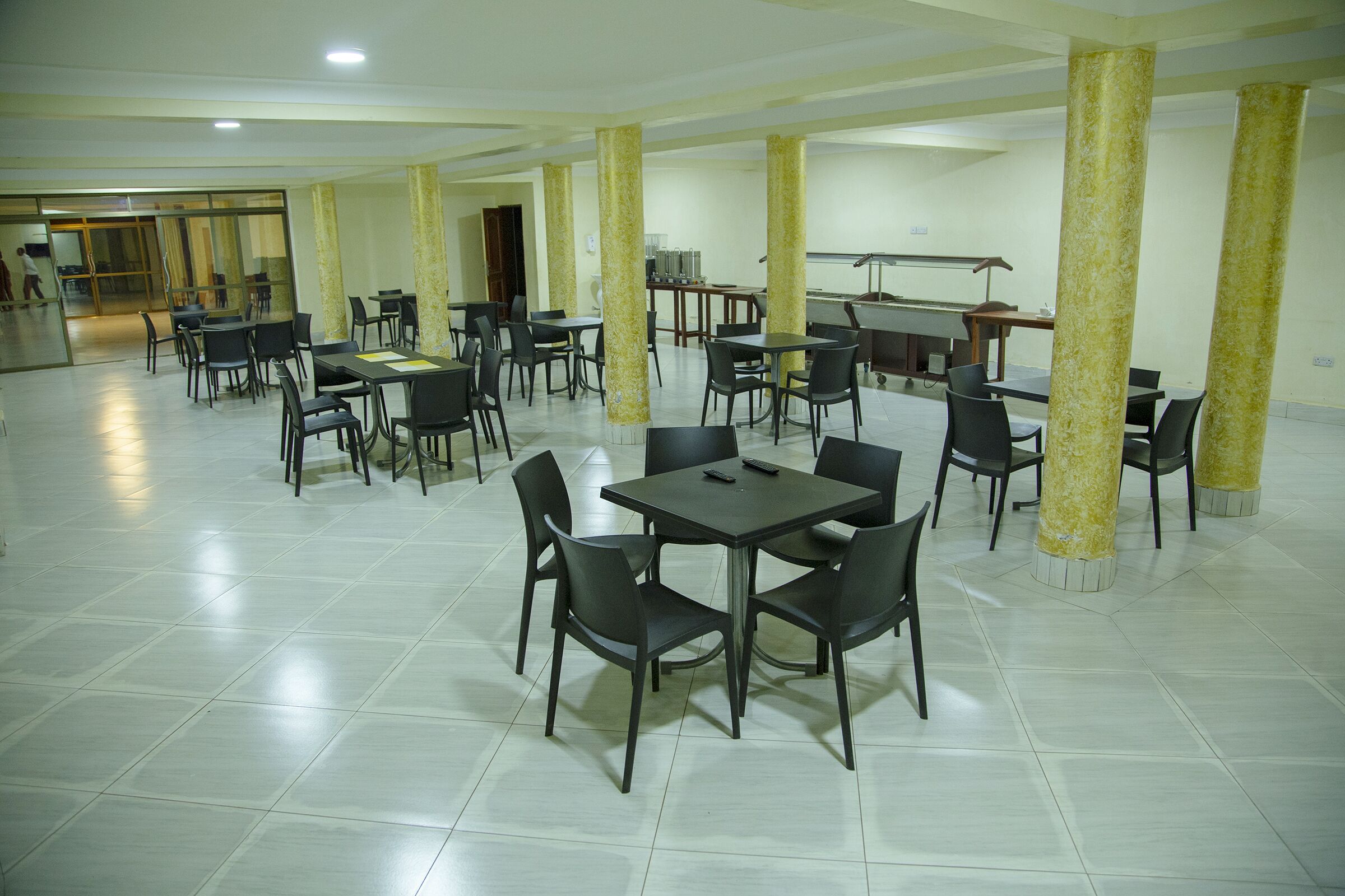 Un vaste espace dédié à la restauration, avec de nombreuses tables et chaises, une scène dotée d’un pupitre, et un grand espace ouvert avec une entrée à porte vitrée.