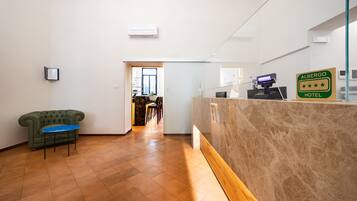 A hotel reception area with a reception desk, a green sofa, and a dining area in the background.