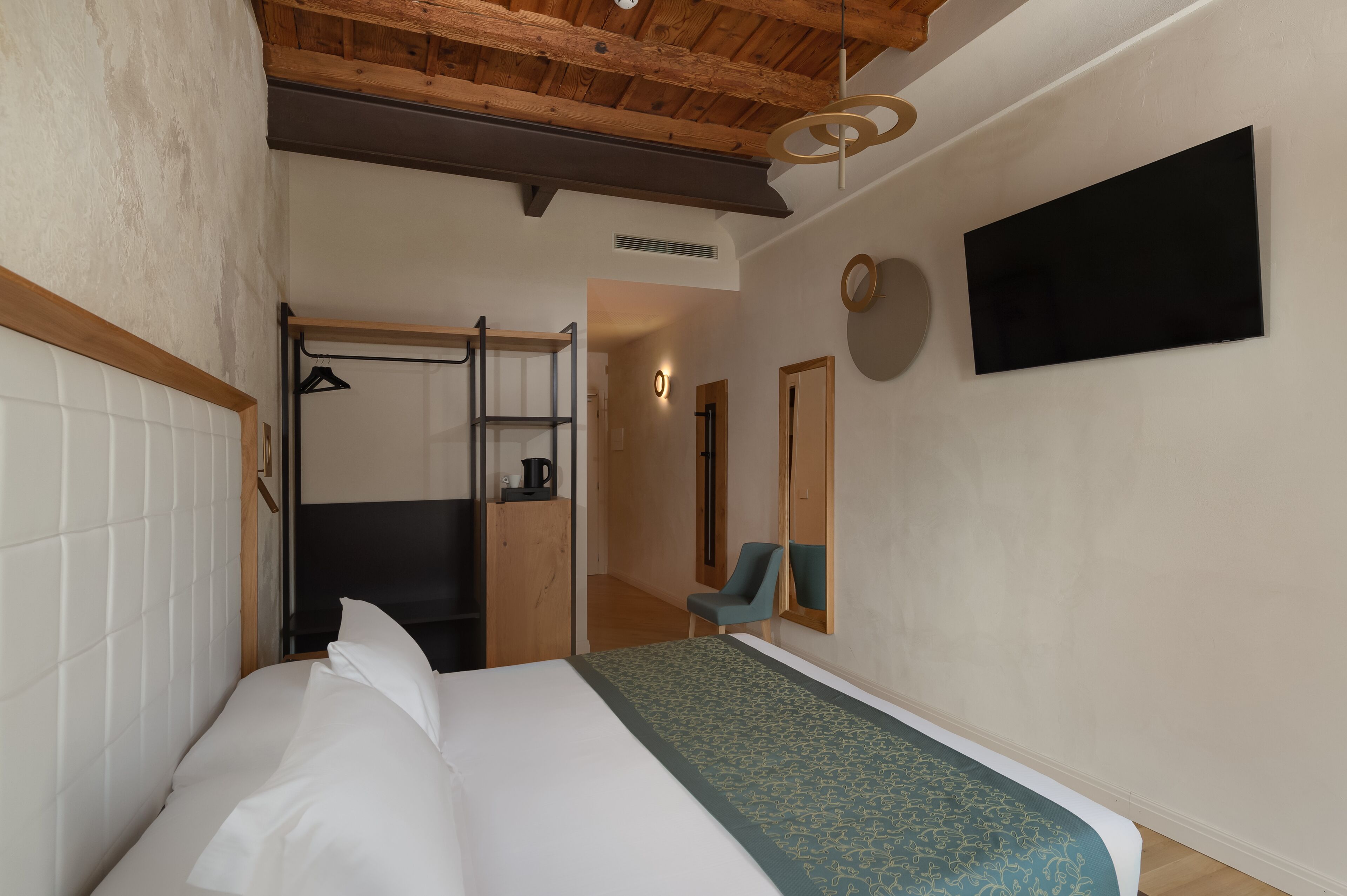 A hotel room with a bed, a TV mounted on the wall, a chair, and a wardrobe with a mirror.