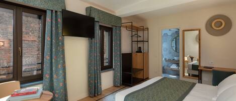 A hotel room with a bed, a TV, a wooden wardrobe, a mirror, and a window with curtains.