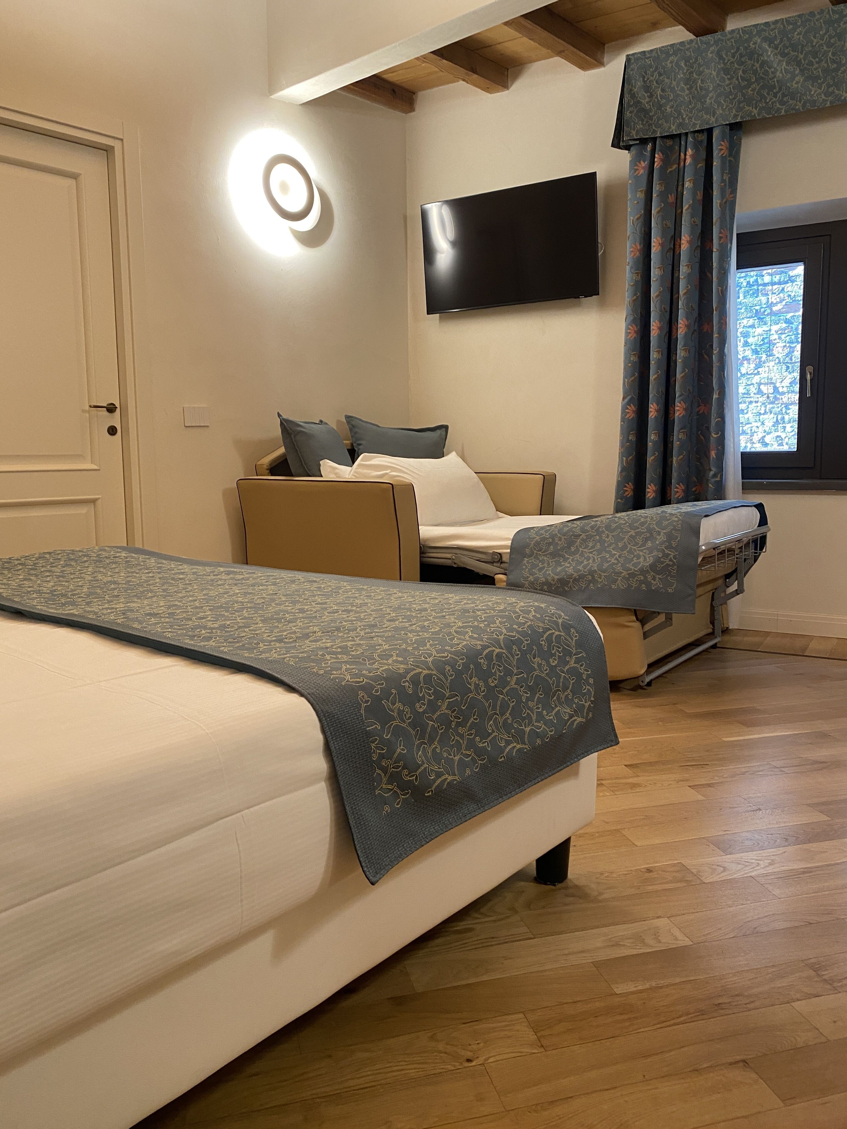 A hotel room with a large bed, a smaller bed, a TV mounted on the wall, and a window with curtains.