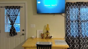 Une pièce comprenant une table à manger en bois, deux chaises noires, un téléviseur fixé au mur affichant « Hisense Roku TV », une petite lampe de table et un panneau indiquant les « règles de la cabane ».