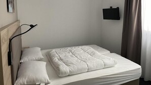 A neatly made bed with white bedding, a wall-mounted lamp, and a flat-screen TV on the wall.