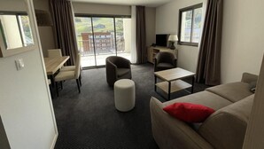 A modern hotel room with a sofa, armchair, ottoman, and a dining table with chairs. There is a TV on a stand, a window with curtains, and a view of the outdoors.