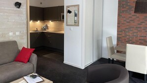 A modern living room with a grey sofa, a red pillow, and a black ottoman. There is a kitchen visible in the background with dark cabinets and a white countertop.