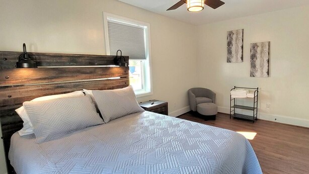 A bedroom with a bed, a nightstand, a chair, a window with blinds, and artwork on the wall. - The Traveler on Main - Suite #6- Across the Street from Back of the Dragon! (Tazewell)