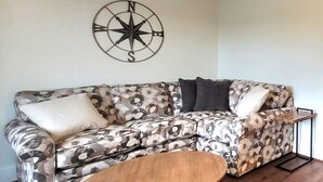 A corner living room with a patterned sofa, a round wooden coffee table, and a wall-mounted compass decoration.