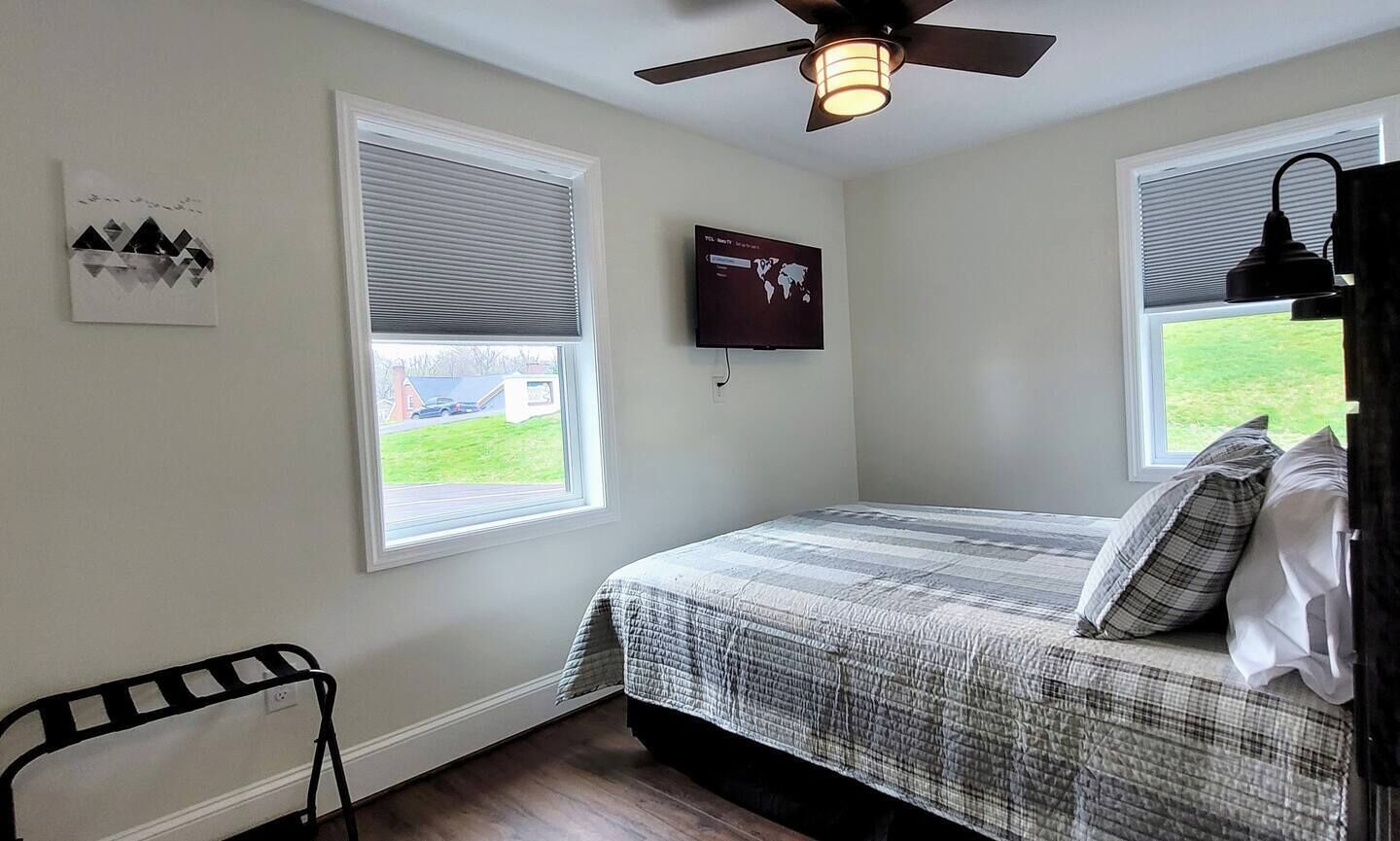 Une chambre à coucher comprenant un lit, deux fenêtres équipées de stores, un ventilateur de plafond, un téléviseur fixé au mur et une petite chaise.