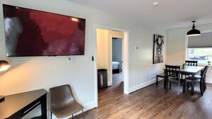 A modern living room with a flat-screen TV, a desk with a lamp, a dining area with a table and chairs, and a hallway leading to another room.