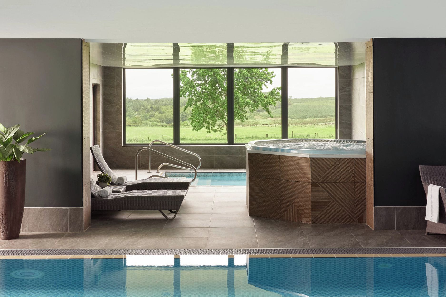 Uma área interna de piscina moderna, com hidromassagem, espreguiçadeiras e vista para uma paisagem verde através de amplas janelas.