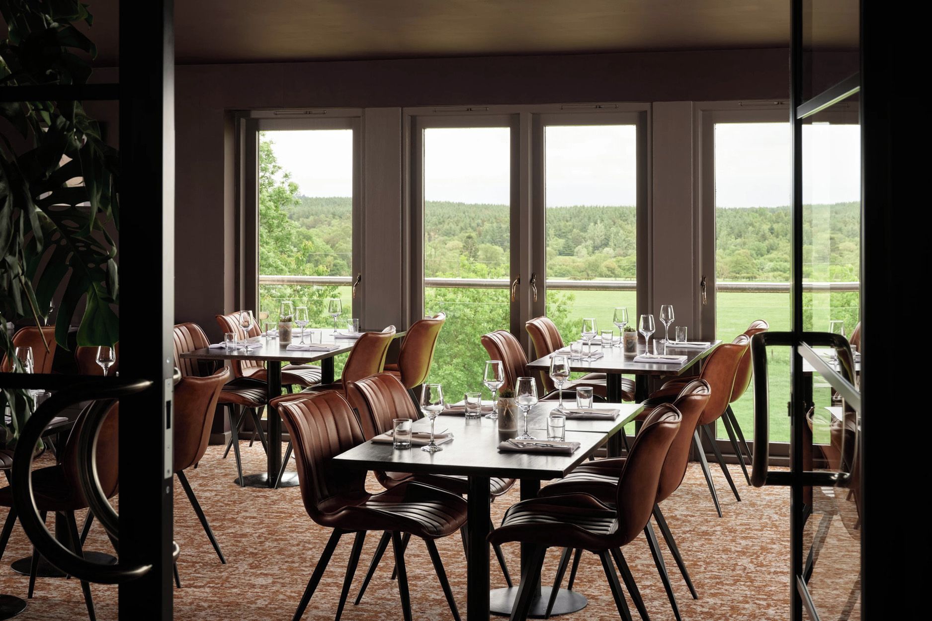 A dining area with tables and chairs, large windows, and a view of greenery.
