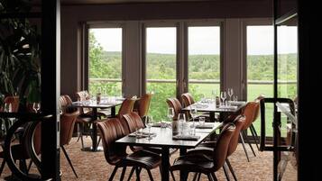 A dining area with tables and chairs, large windows, and a view of greenery.
