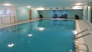 Una piscina interior con suelo de baldosas, zona de vestuarios y área de descanso con sillas.