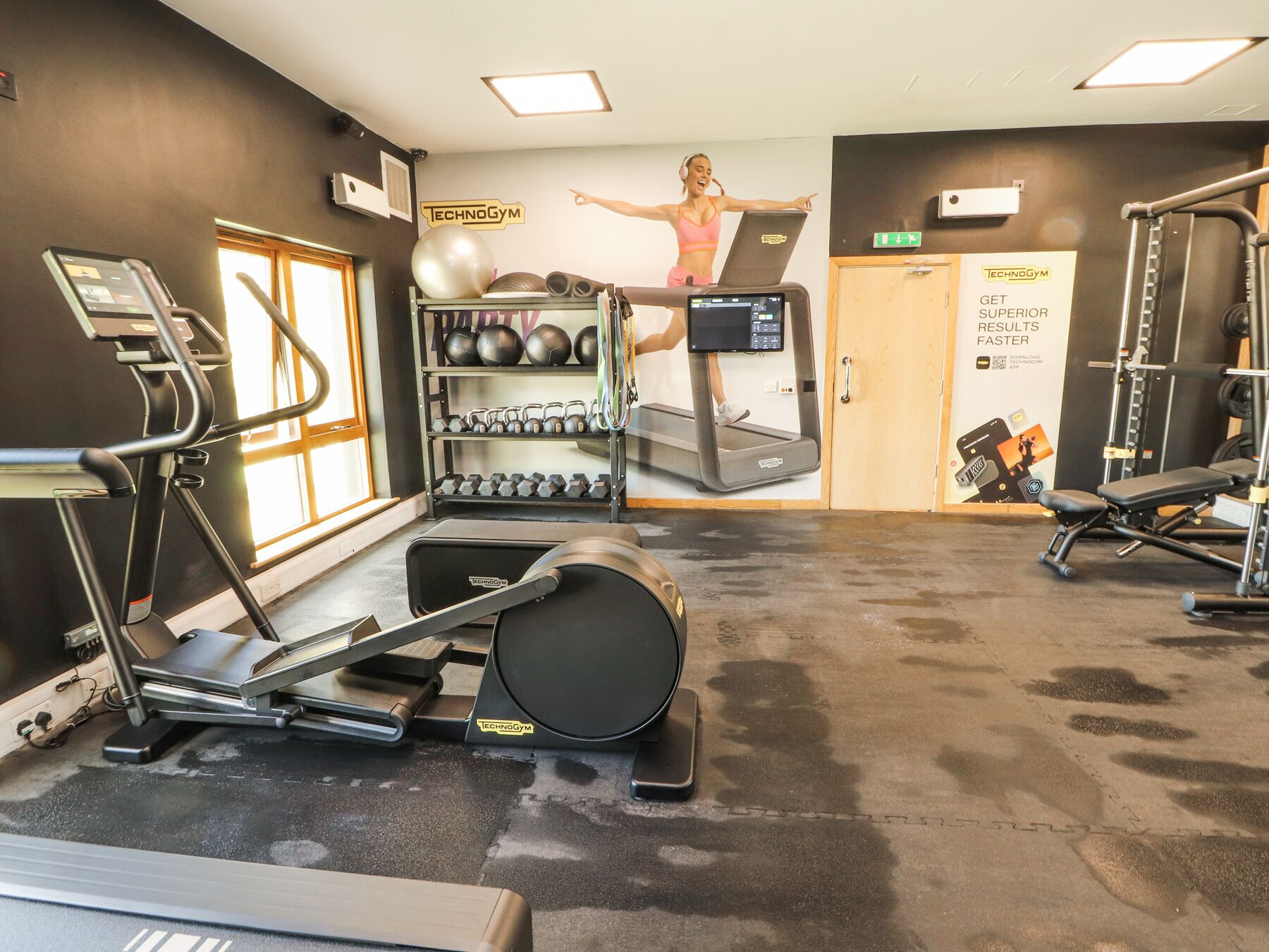 A gym with treadmills, a wall-mounted elliptical, free weights, and a poster of a woman exercising.