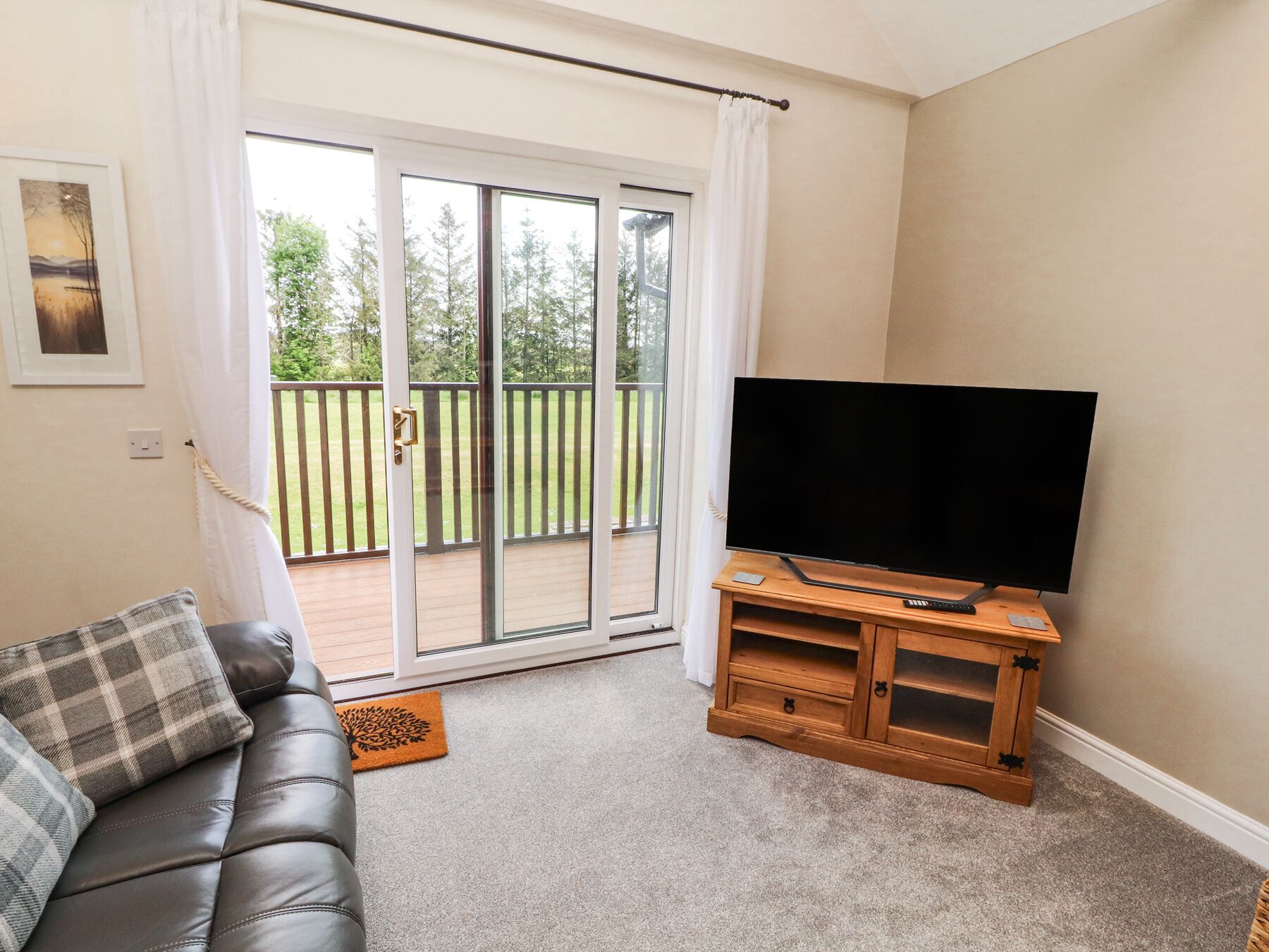 A living room with a leather couch, a flat-screen TV on a wooden stand, and a sliding glass door leading to a balcony with a wooden railing.