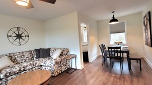 A living room with a floral-patterned sofa, a wooden coffee table, and a dining area with a table and chairs. A compass wall decoration is visible, and there is a window providing natural light. - The Traveler on Main - Suite #4- Across the Street from Back of the Dragon! (Tazewell)