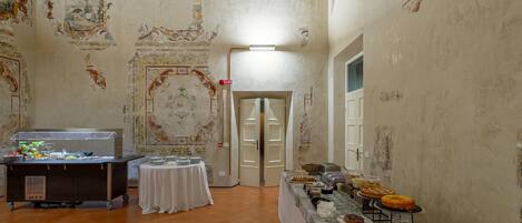 A room with a buffet setup, a small round table with a white tablecloth, and a wall with decorative frescoes.