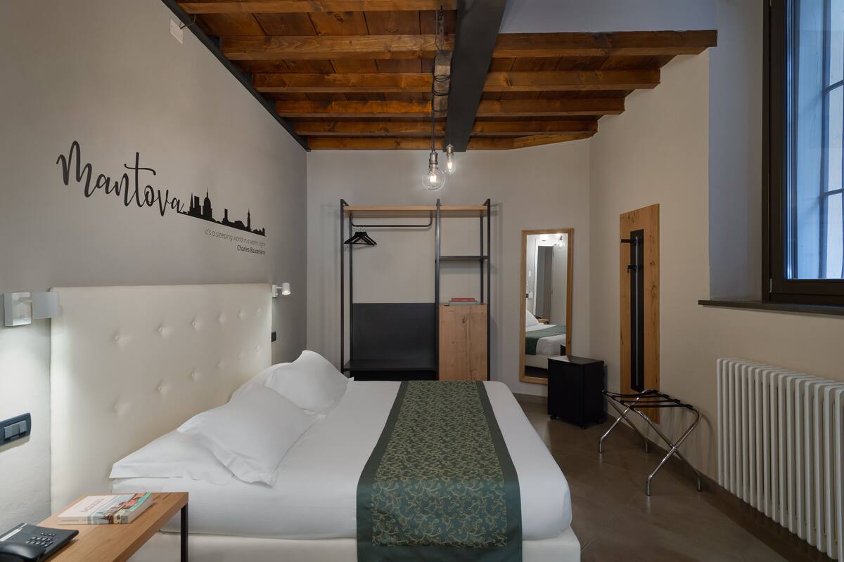 Una habitación de hotel con cama, mesita de noche, lámpara colgante, radiador y ventana.