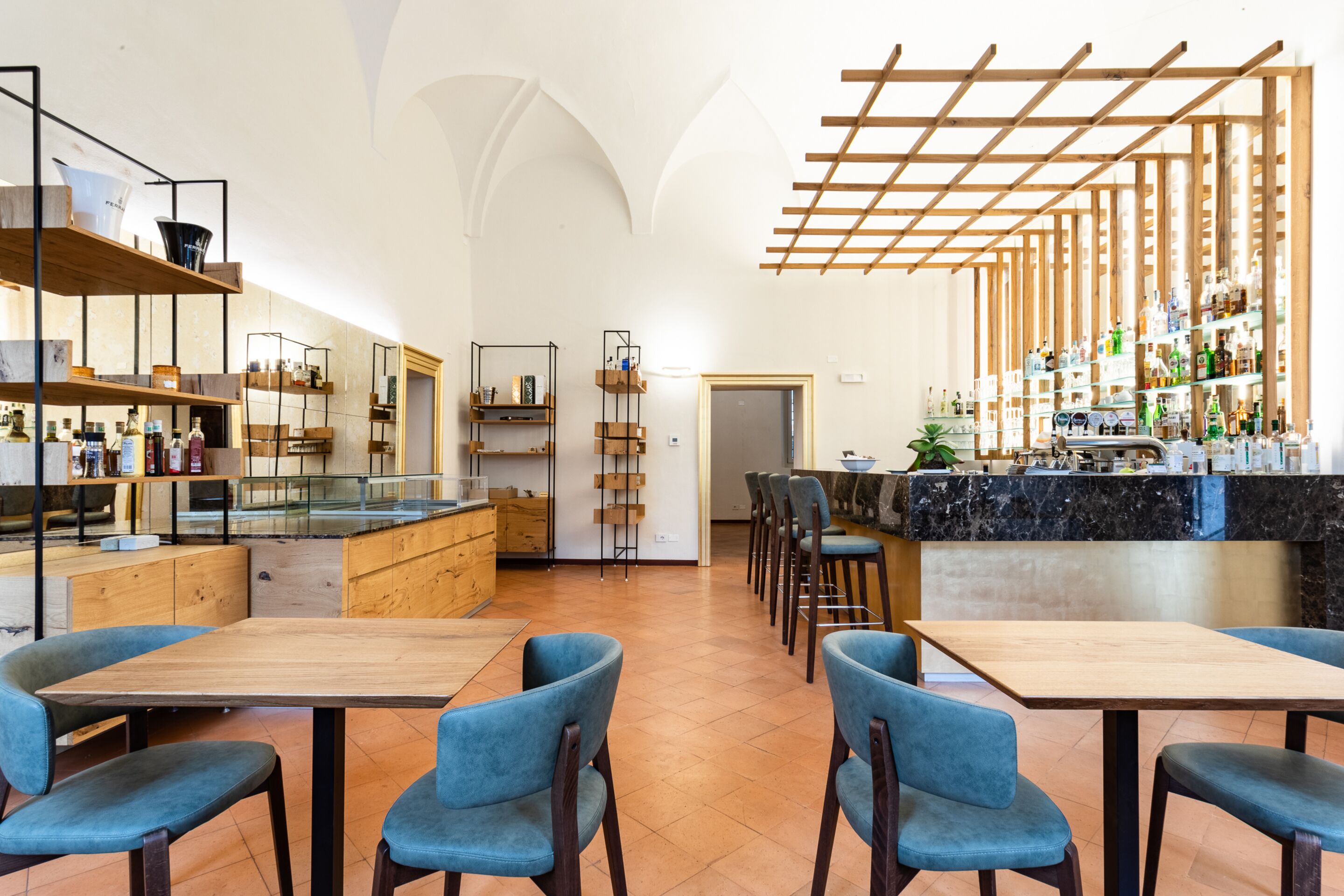 A modern restaurant with wooden tables and blue chairs, a bar area with shelves of bottles, and a high ceiling with wooden beams.