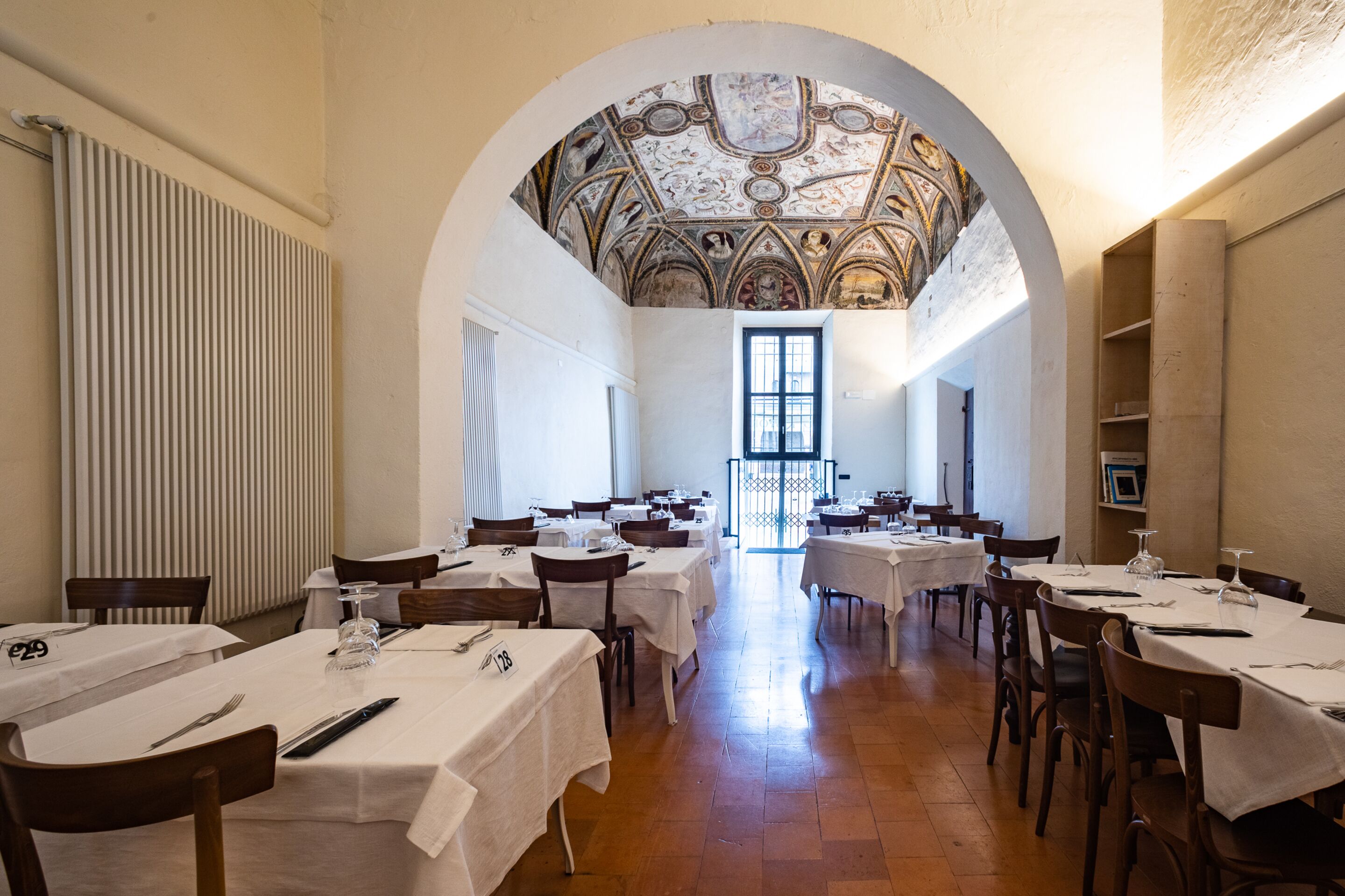 A dining area with tables set for service, a high arched ceiling with frescoes, and a view through a doorway.