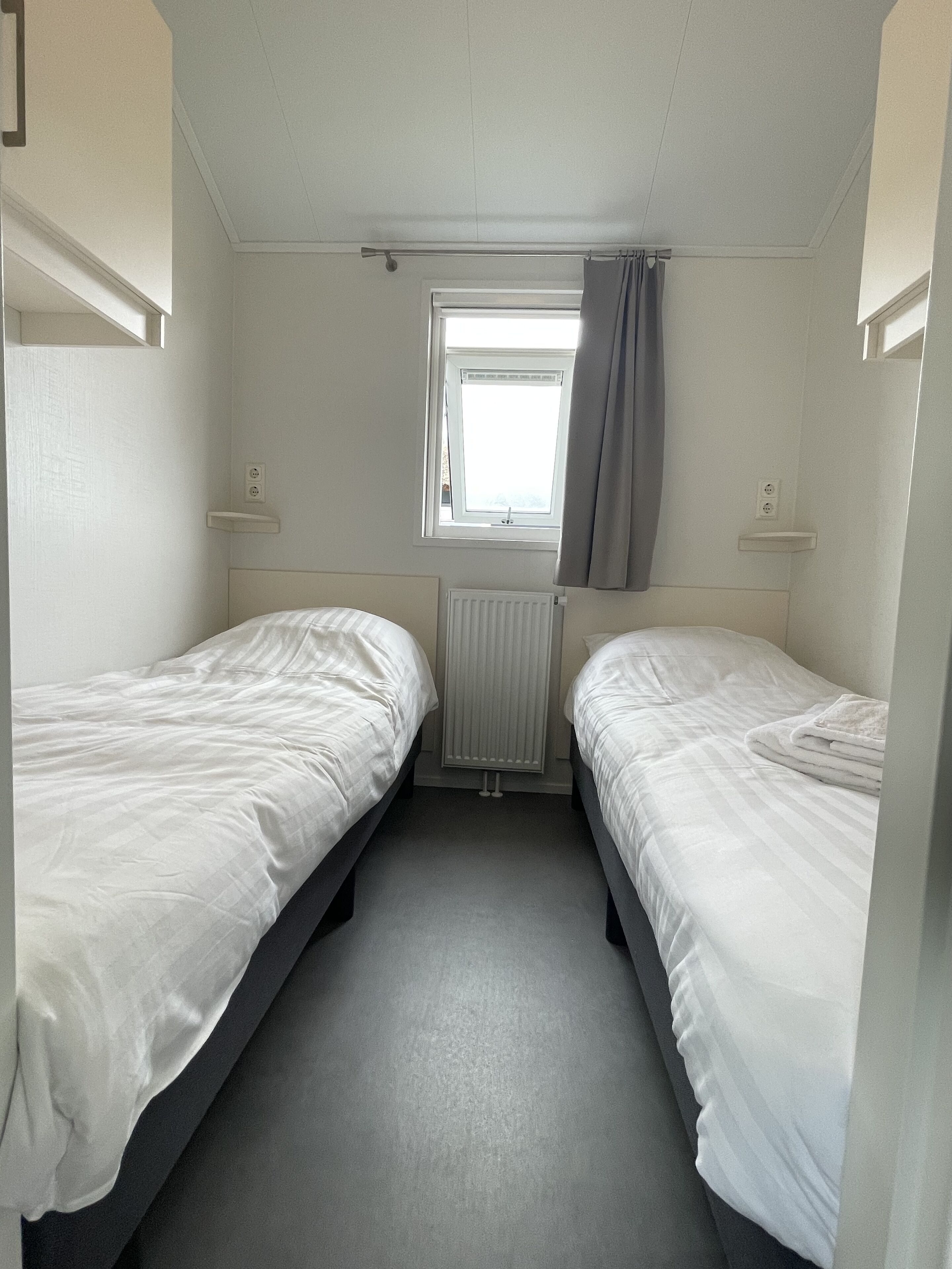 A small, single-room accommodation with two single beds, a window with curtains, and a radiator.