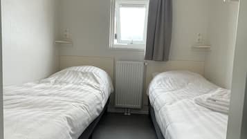 Un petit logement composé d’une seule pièce, comprenant deux lits simples, une fenêtre avec des rideaux et un radiateur.