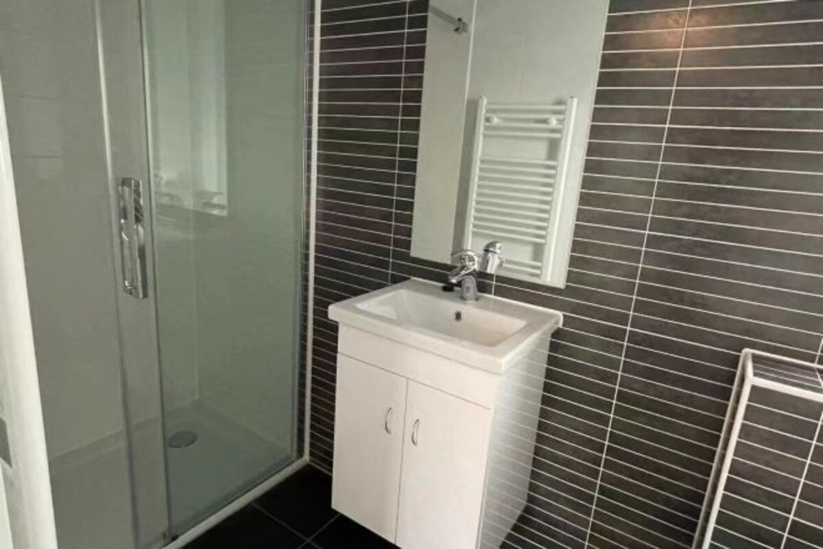 Un baño con cabina de ducha de vidrio, un lavabo blanco con espejo y paredes de azulejos oscuros.