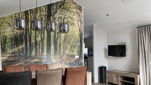 Una sala de estar moderna con un gran mural de un bosque, una zona de comedor con sillas marrones, un televisor montado en la pared y un piso de madera con estampado de espiga.