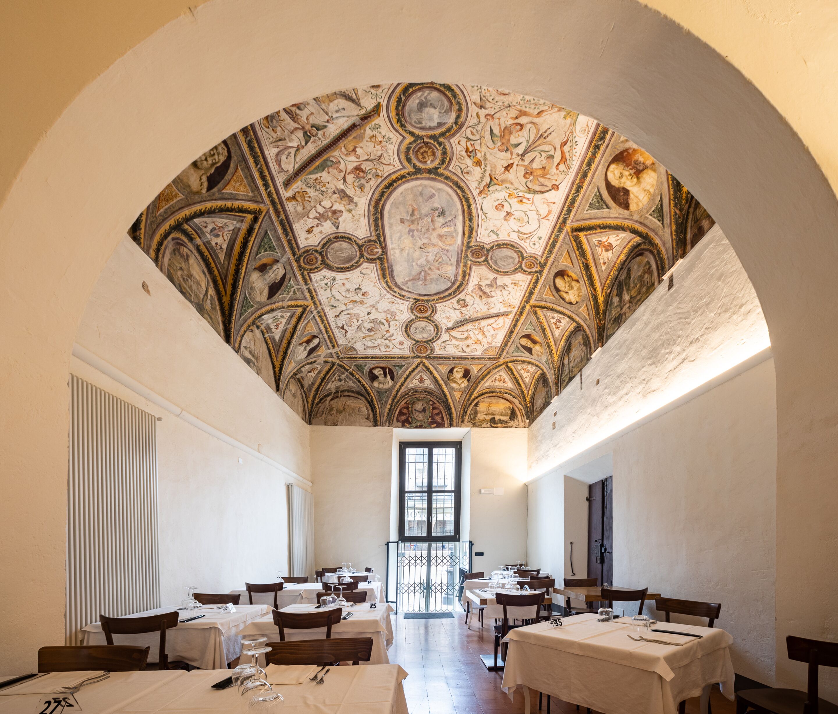 A dining area with tables and chairs, a high ceiling with intricate frescoes, and a large window at the far end.