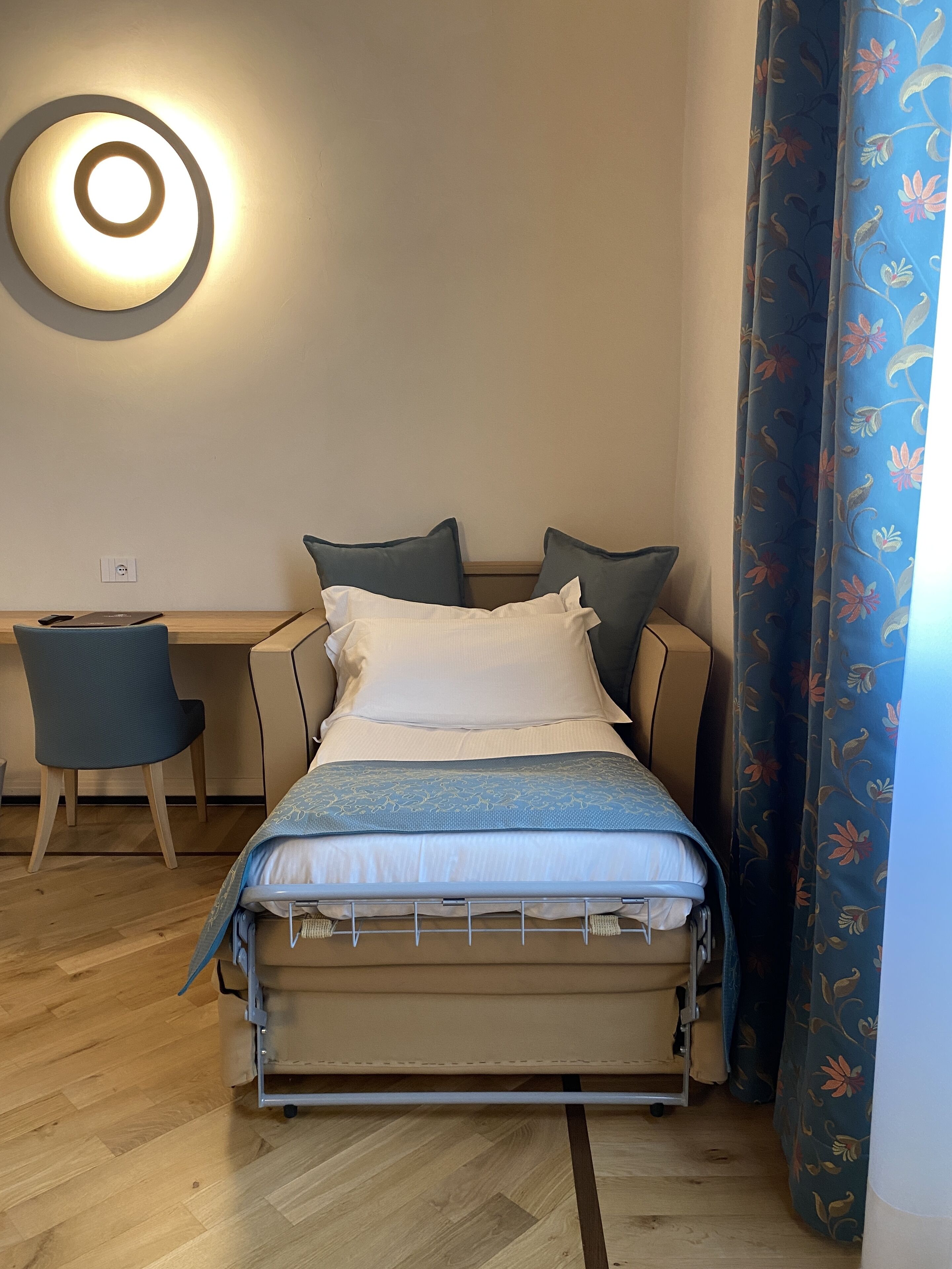 Uma cama compacta com estrutura de metal, uma mesa de cabeceira com luminária e uma escrivaninha com cadeira.