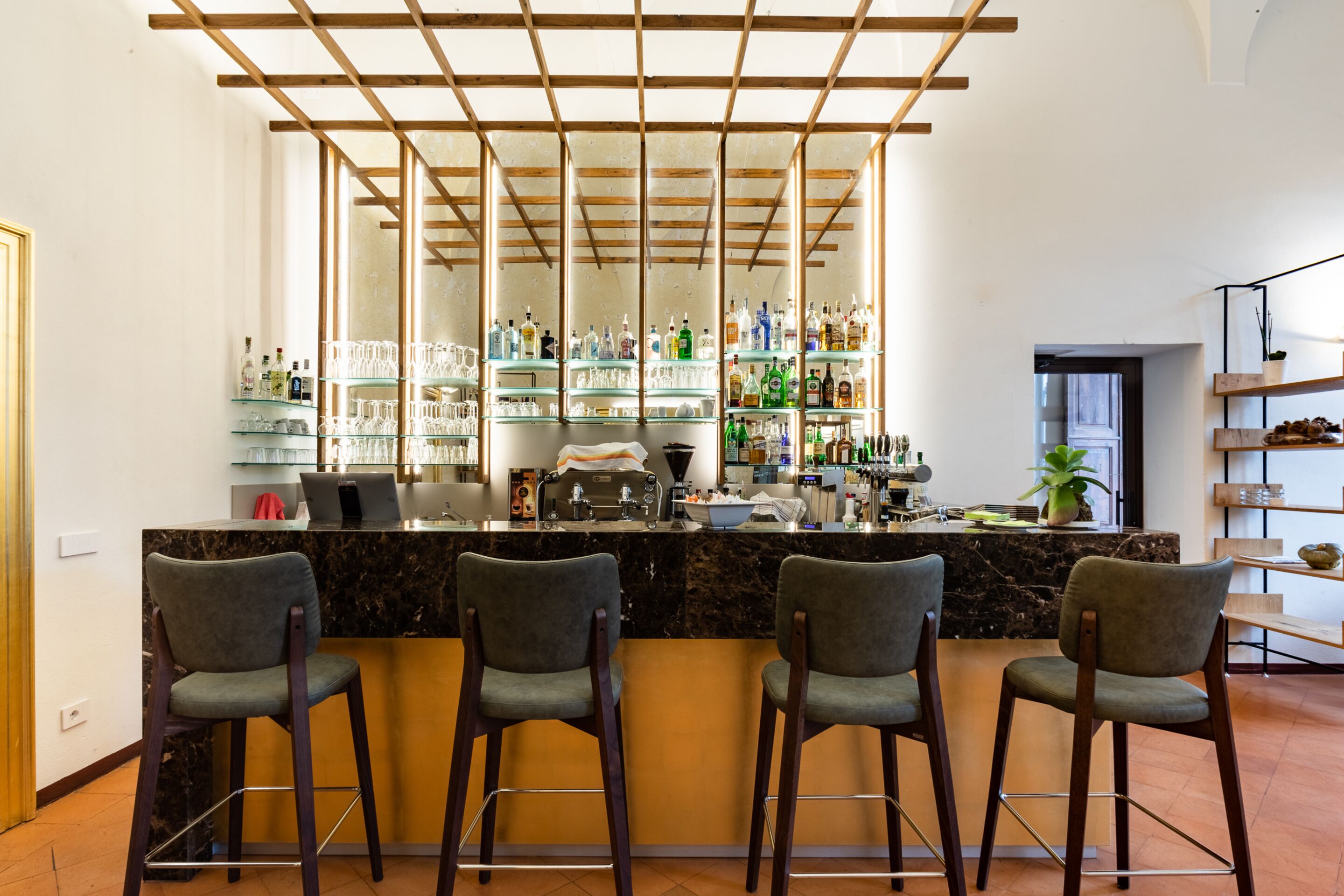 Eine Bar mit einer Marmorplatte, drei Barhocker und Regalen voller Flaschen und Gläsern.