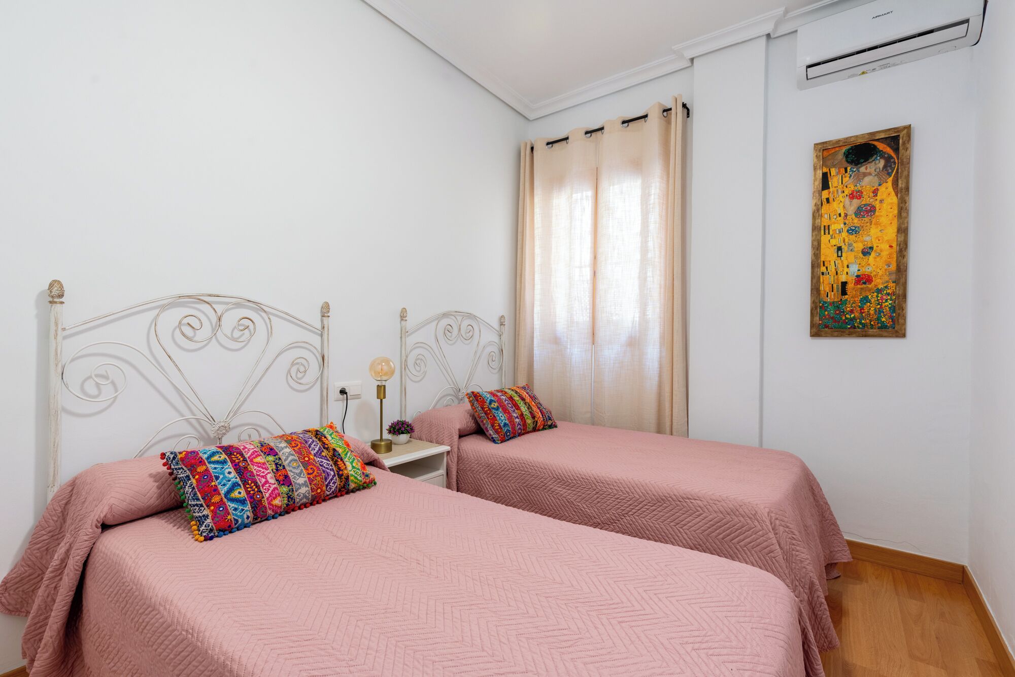 A bedroom with two beds, a nightstand, a lamp, a painting, and a window with curtains.