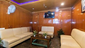A room with wooden panel walls, a flat-screen TV mounted on the wall, two beige sofas, a glass coffee table, and a standing fan. - Hotel Telehaus Lalbagh Botanical Garden (Bengaluru)
