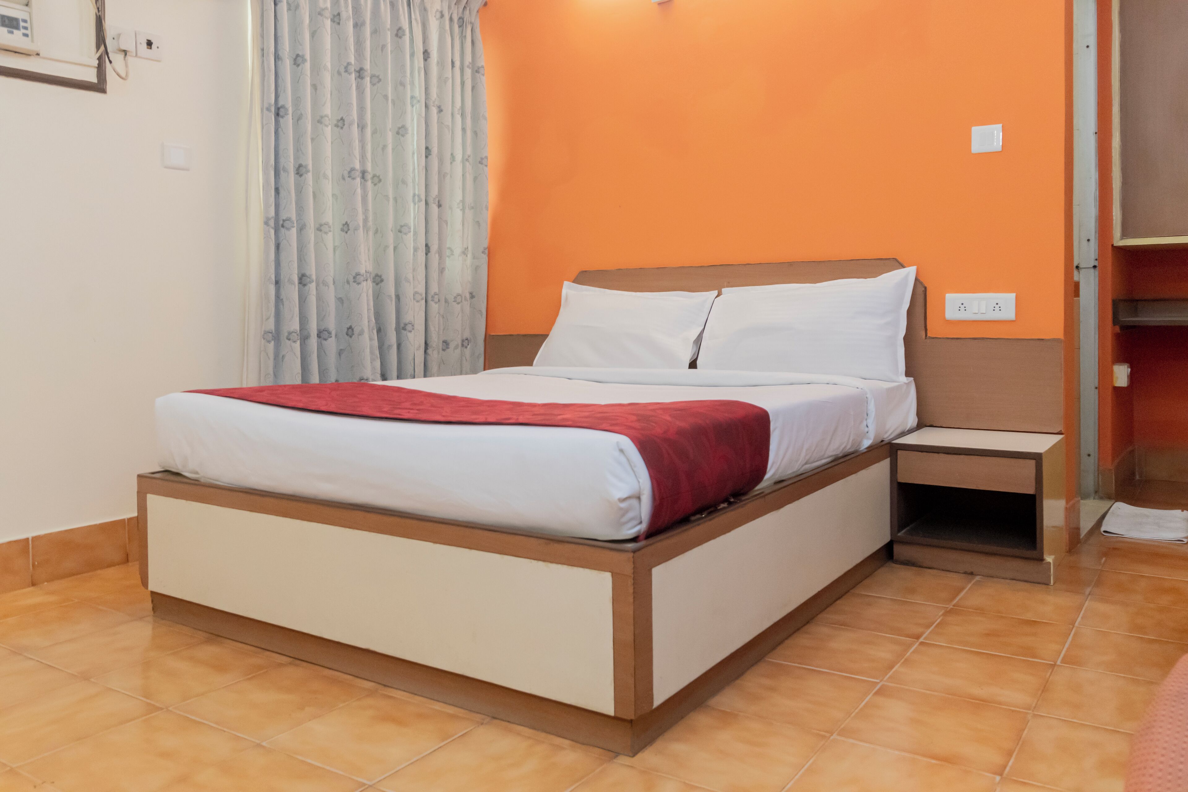 A single bed with a wooden frame, white bedding, and a red blanket. There is a nightstand with a lamp and a power outlet. The room has orange walls and a window with patterned curtains.
