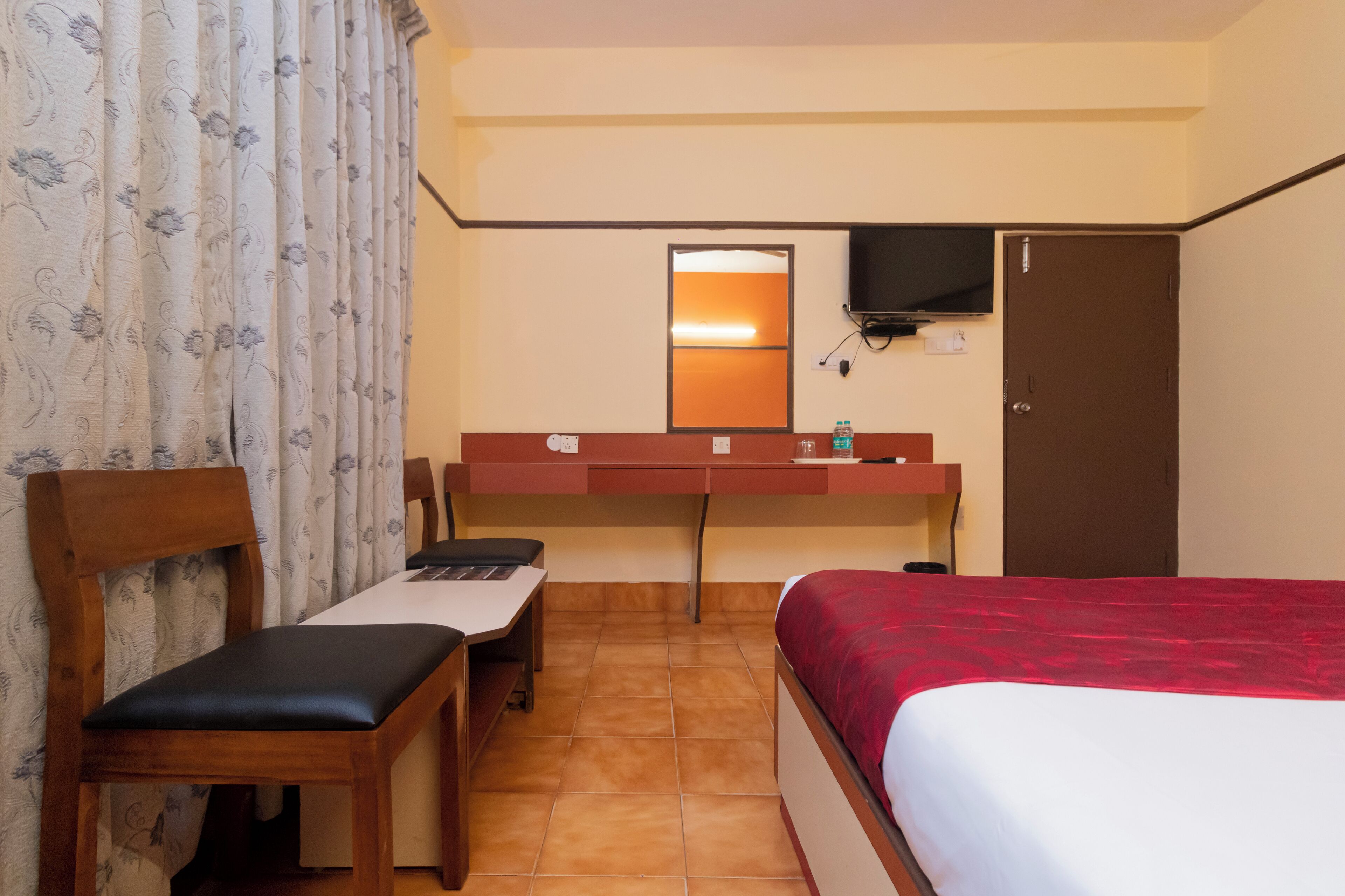 A hotel room with a bed, a desk with a chair, a television mounted on the wall, and a door.